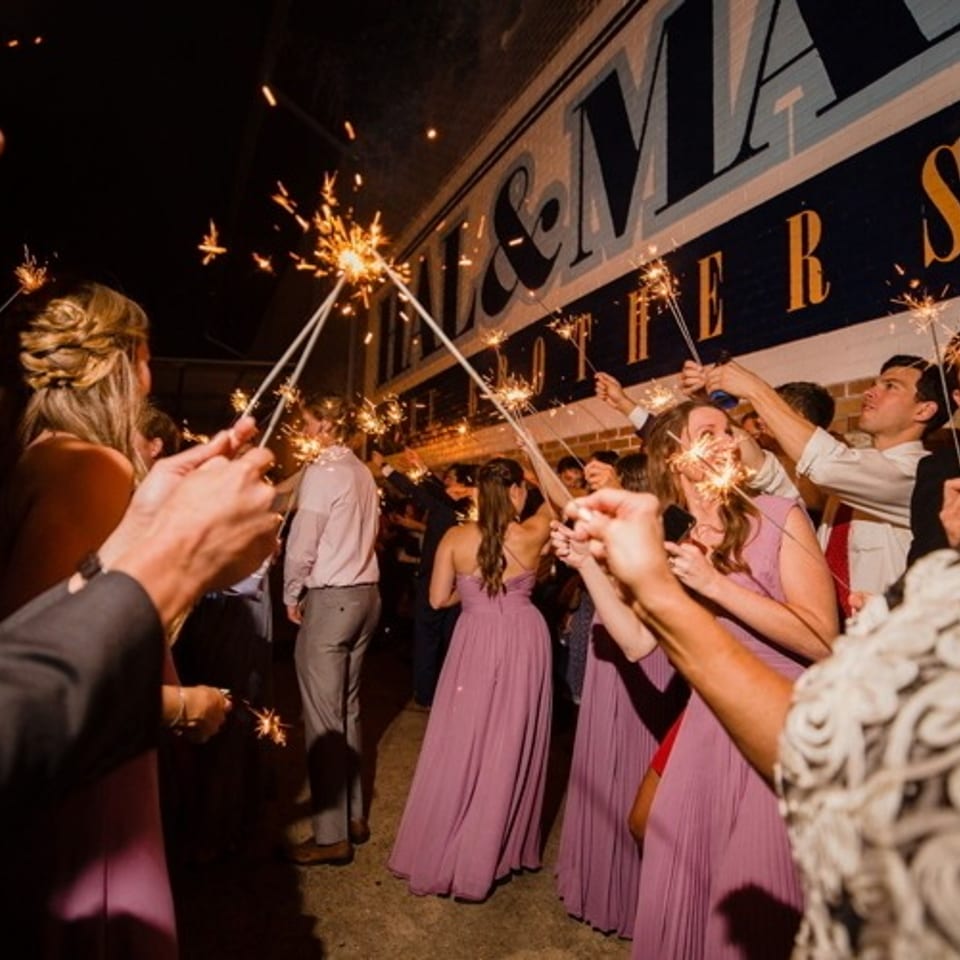 Wedding guests raise sparkling sparklers beneath a large vintage theater-style sign during an evening celebration.