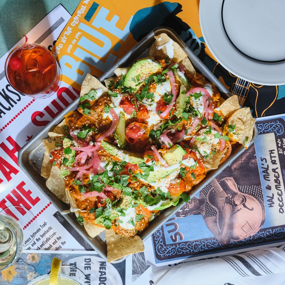 Loaded nachos topped with avocado, tomatoes, pickled onions, cilantro, sesame seeds, and creamy sauce beside colorful cocktails and a plate.