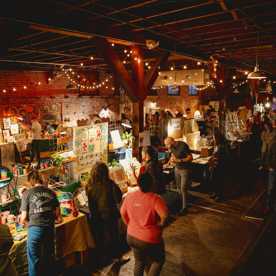 Crowded brick-walled market hall with vendors displaying handmade crafts beneath warm string lights.