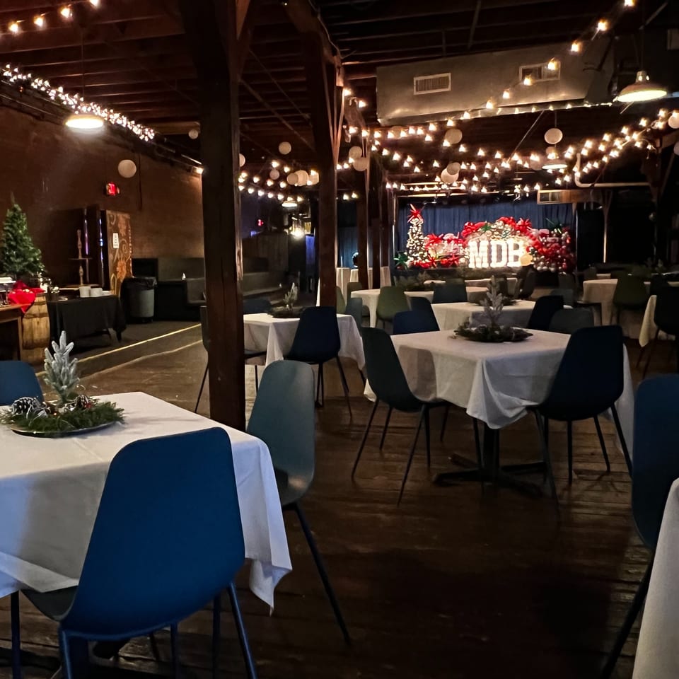 Festive restaurant dining room with white tablecloths, blue chairs, glowing string lights, and a holiday floral stage display.