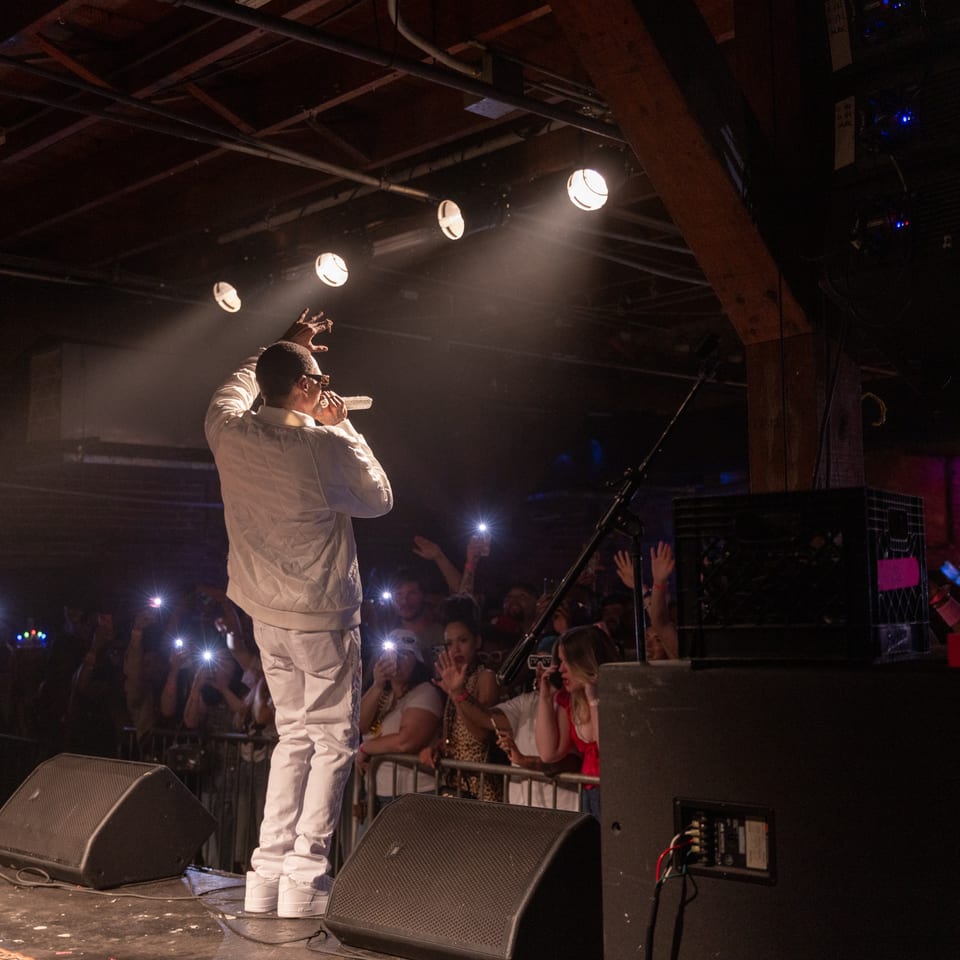 Performer in white clothing sings onstage beneath spotlights as an enthusiastic crowd raises hands and phone cameras.
