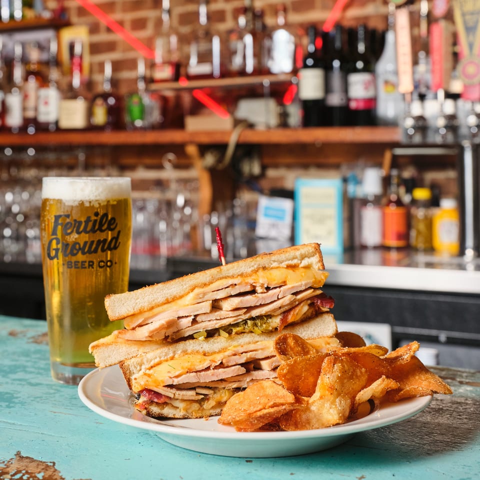 Stacked grilled sandwiches with melted cheese and potato chips beside a glass of beer at a colorful bar.