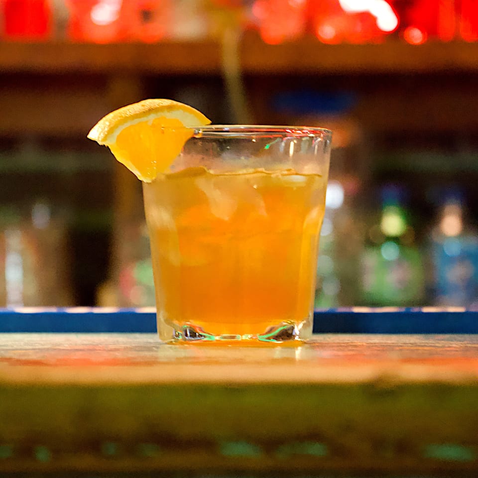 Orange cocktail in a rocks glass garnished with an orange wedge, set against colorful blurred bar lights.