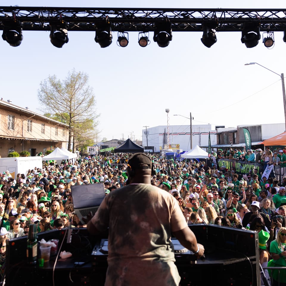 DJ performs onstage before a large outdoor crowd wearing green at a lively festival with tents and Jameson banners.
