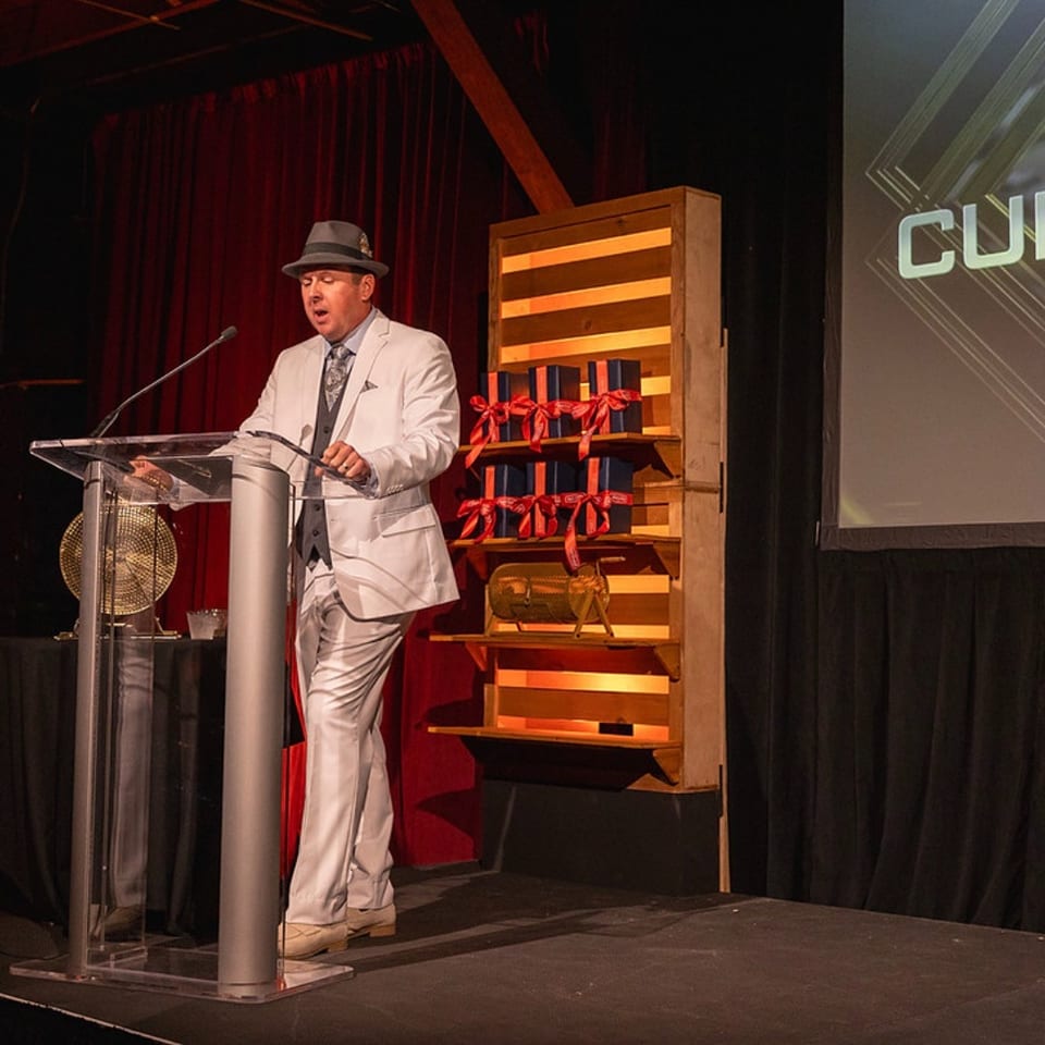 Man in a white suit and fedora speaks at a podium on a theater stage beside a screen displaying cultural event text.