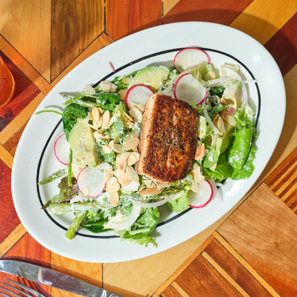 Grilled salmon fillet atop a leafy green salad with avocado, radishes, onions, asparagus, and sliced almonds on a restaurant table.