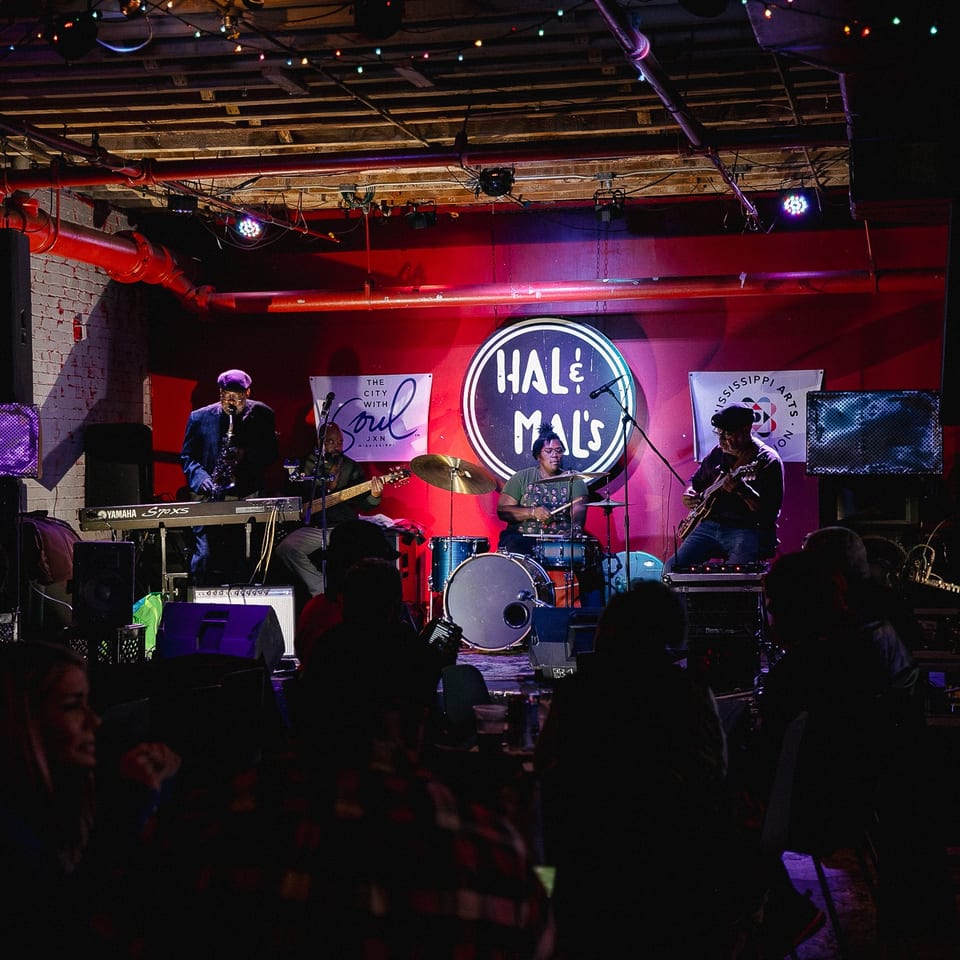 Live band performs onstage at Half Shell’s, with saxophone, keyboard, drums, and guitar under colorful lights before a seated audience.
