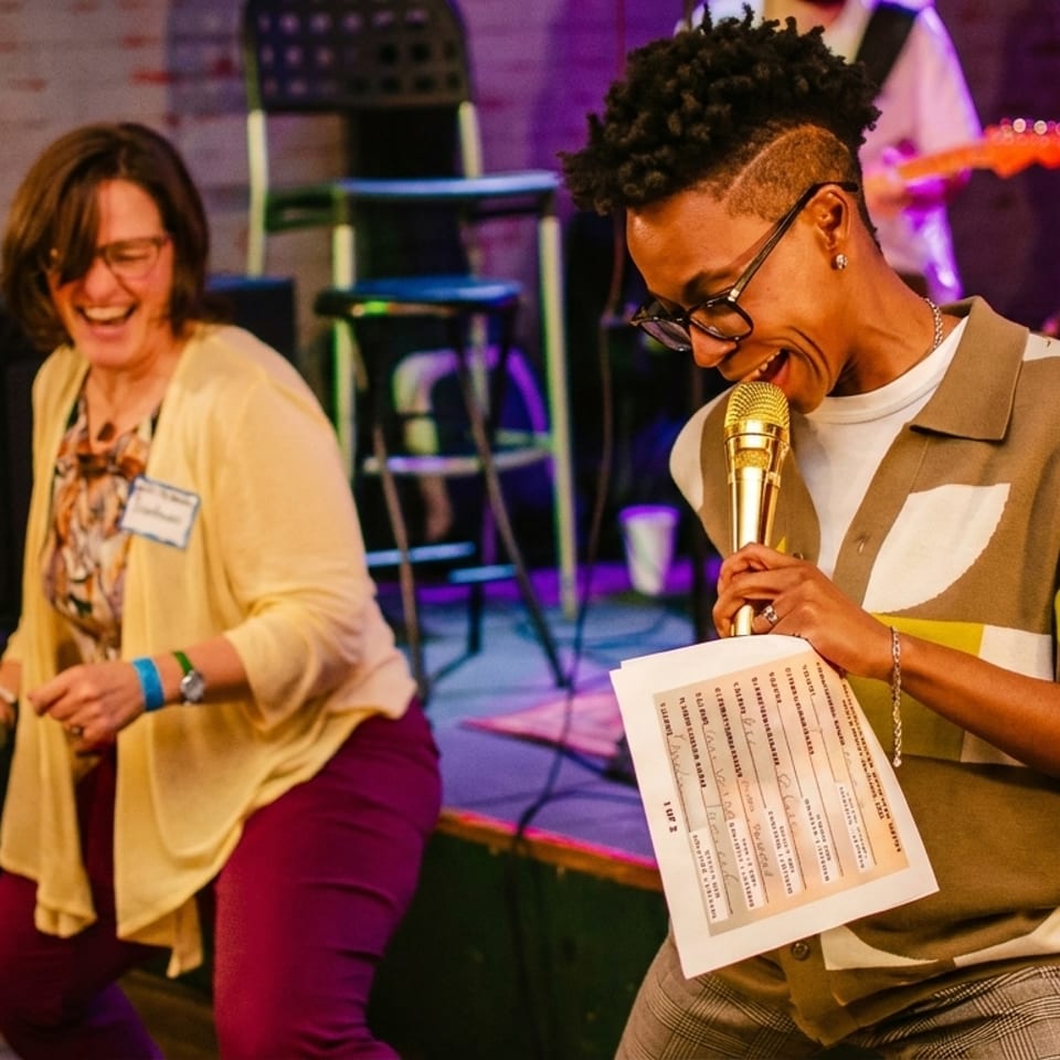 Two people laughing and dancing on stage, one holding a golden microphone and sheet music.