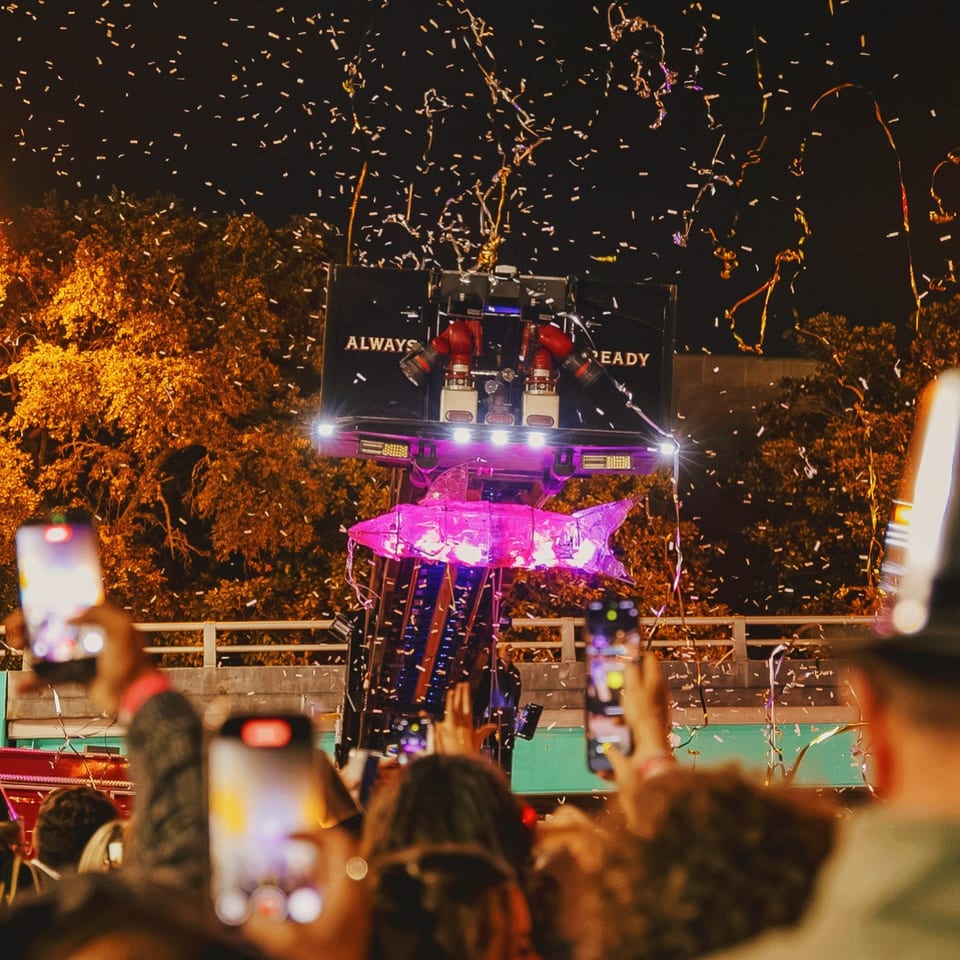 Crowd holds up phones as a brightly lit parade float passes beneath a shower of confetti at night.