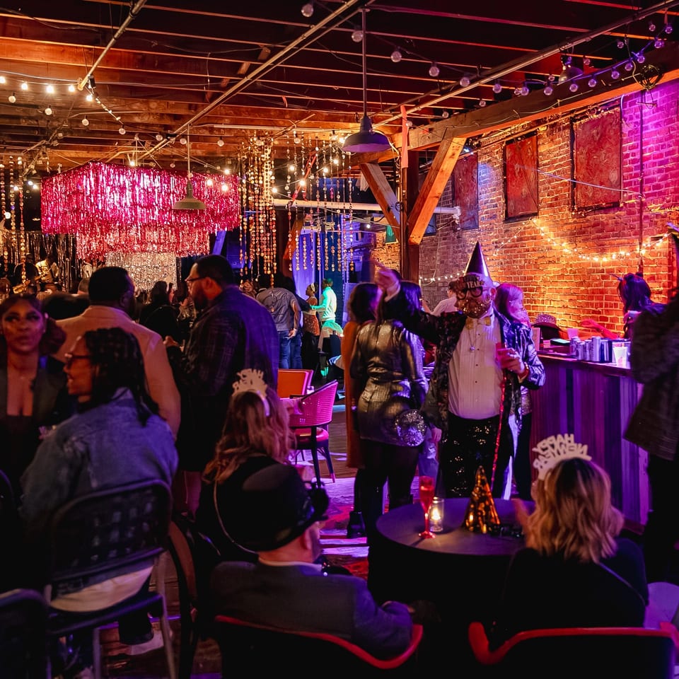 Crowded restaurant nightlife scene with guests in festive costumes, colorful lighting, exposed brick walls, and sparkling decorations.