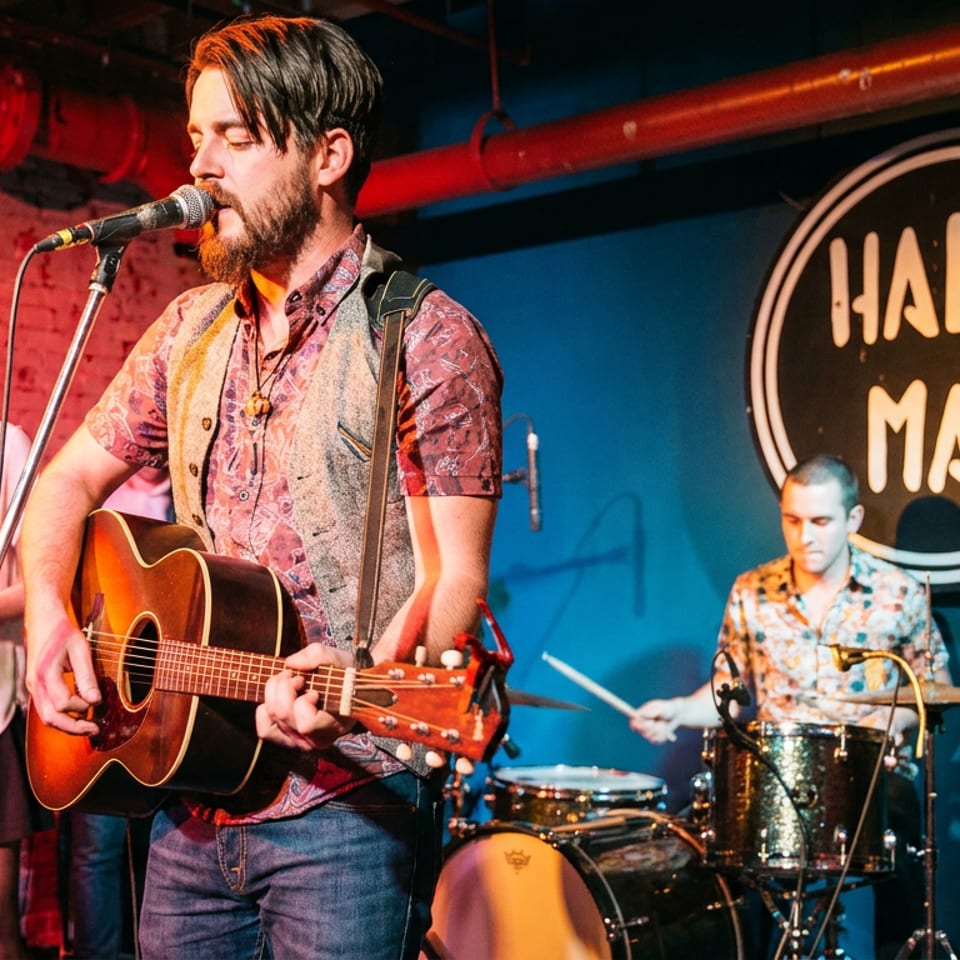 A band performs live on stage at Hal & Mal's, featuring a singer-guitarist, tambourine player, and drummer.