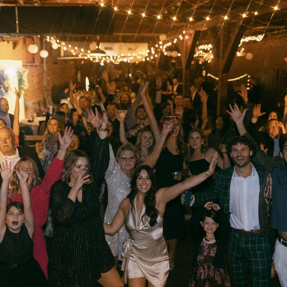 Large crowd celebrating with raised hands inside a warmly lit restaurant or event space decorated with string lights.
