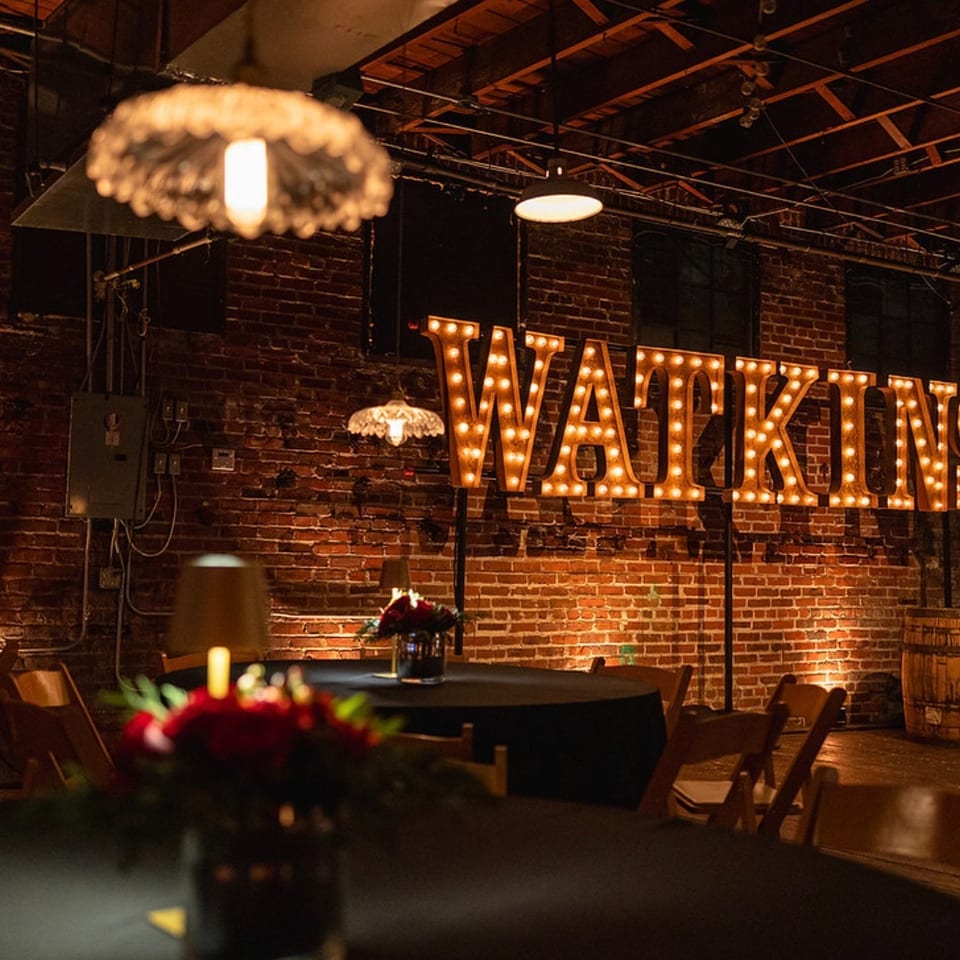 Warmly lit restaurant interior with brick walls, black-clothed tables, floral centerpieces, and a marquee sign reading “MADEKINS.”