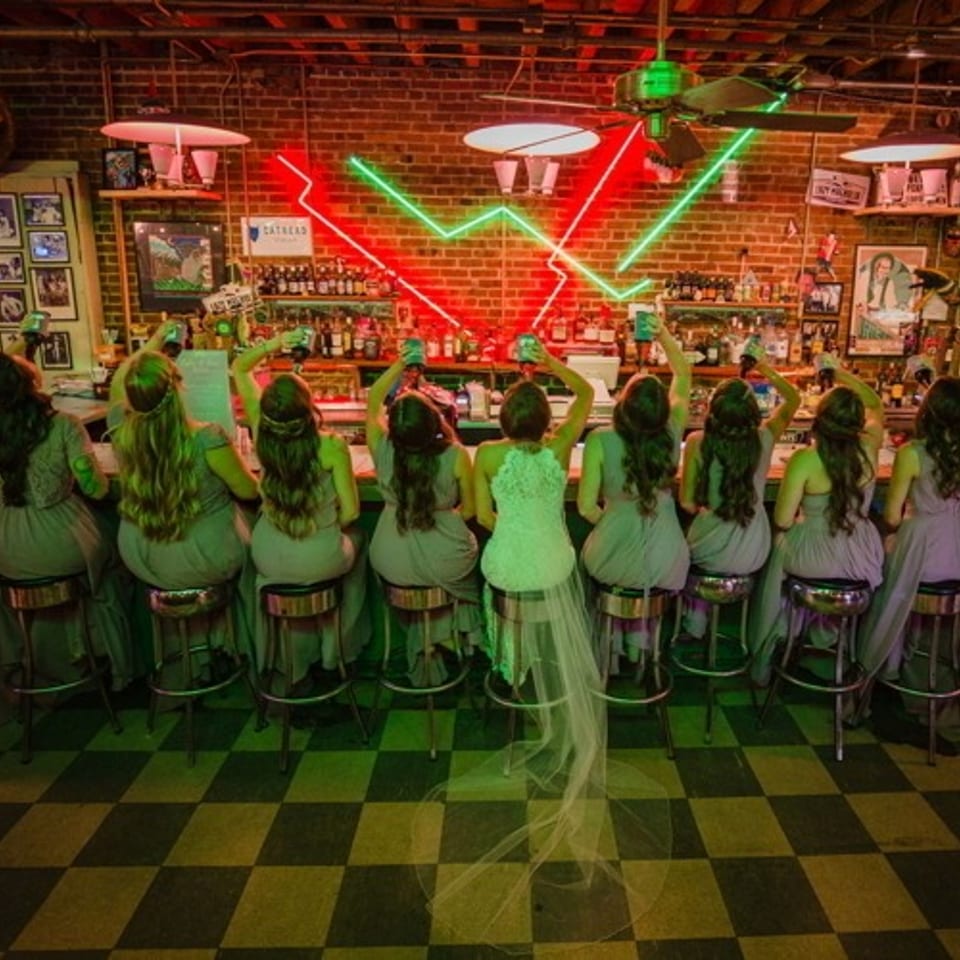 Group of women seated at a neon-lit bar, surrounded by liquor bottles, vintage posters, and a checkered floor.