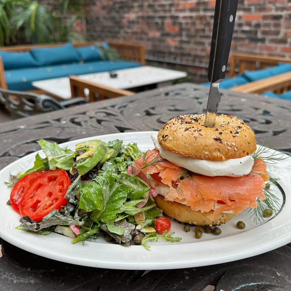 Sesame bagel stacked with smoked salmon, cream cheese, and dill beside a fresh green salad on a restaurant patio table.