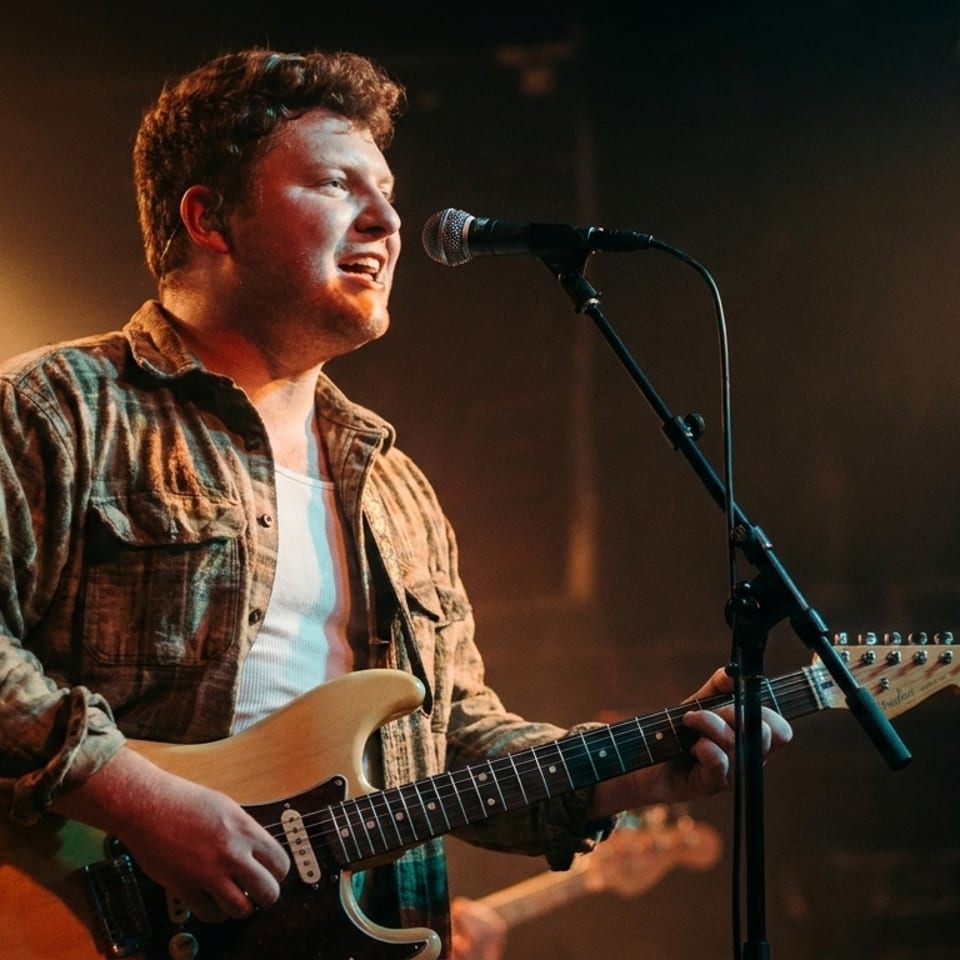 A male musician sings into a microphone while playing an electric guitar on a dimly lit stage.